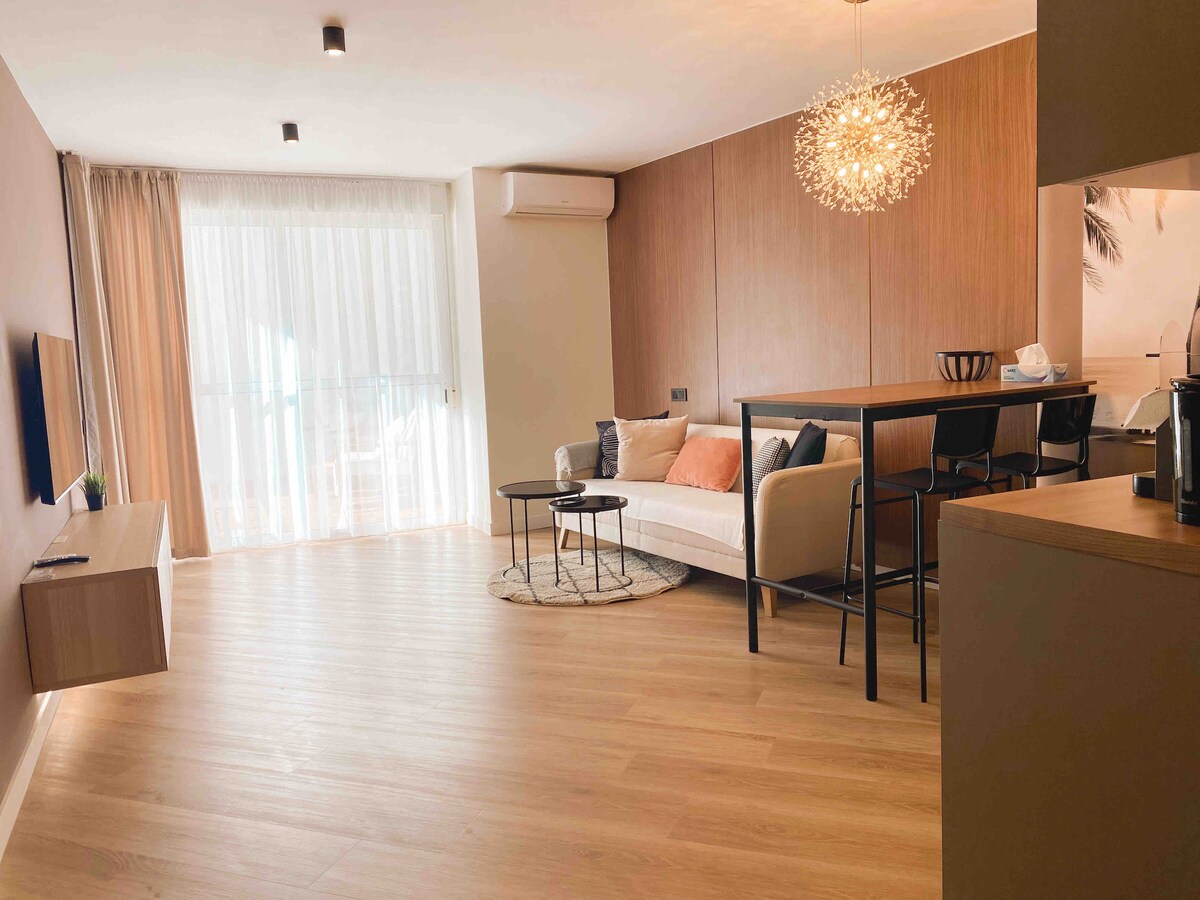 An open-plan living area is shown, featuring a comfortable sofa paired with a circular coffee table. A dining table for two sits nearby, illuminated by a decorative light fixture. Large glass doors bring in natural light and provide access to a balcony with views.