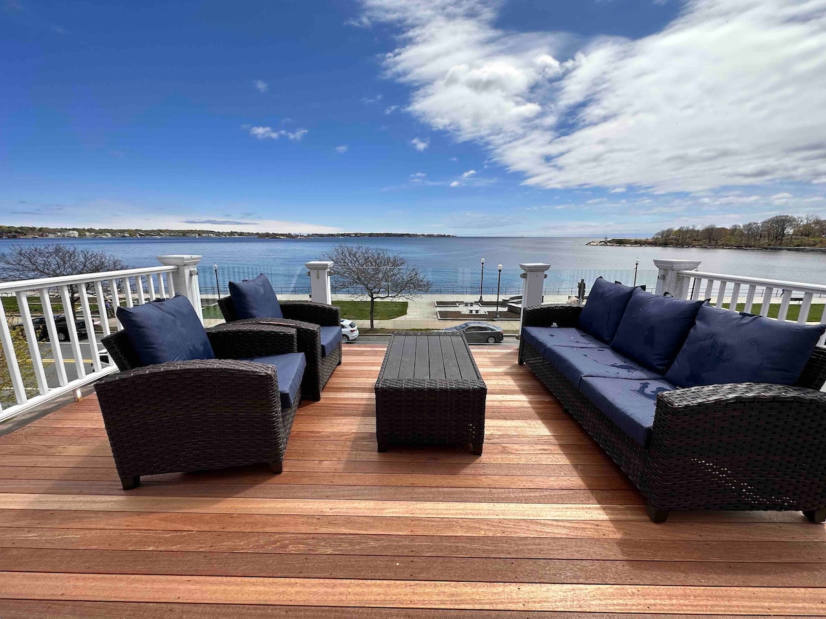 A spacious outdoor seating area features dark wicker furniture with plush navy cushions, arranged around a low table. The view overlooks a body of water, with a clear sky above and nearby trees visible in the background.