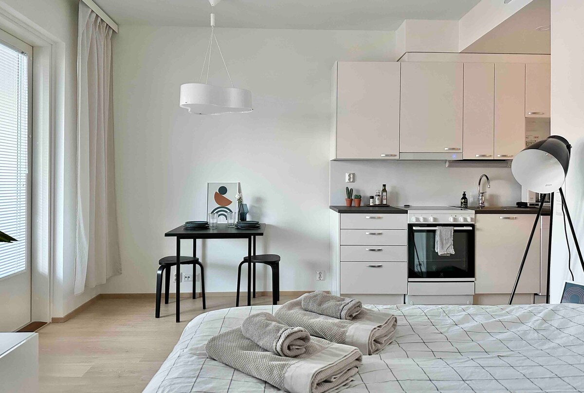 A minimalist studio layout includes a spacious bed adorned with neatly arranged towels. A compact dining table with two chairs is positioned near the kitchenette, which features essential appliances. Abundant natural light enters through a large window, complemented by soft, neutral tones throughout.