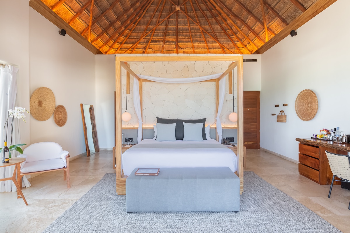 A spacious bedroom is highlighted by a four-poster bed with a light-colored canopy and neutral bedding. Natural materials are used throughout, with a woven ceiling and stone wall providing texture. A seating area is featured, along with decorative mirrors and woven baskets adorning the walls.