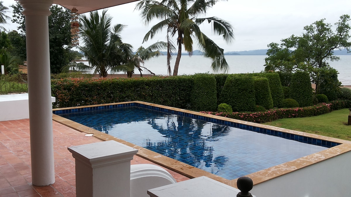 A private swimming pool is bordered by lush greenery and manicured hedges, with palm trees gently framing the serene view of the ocean. The patio area features terracotta tiles, complementing the tranquil setting.
