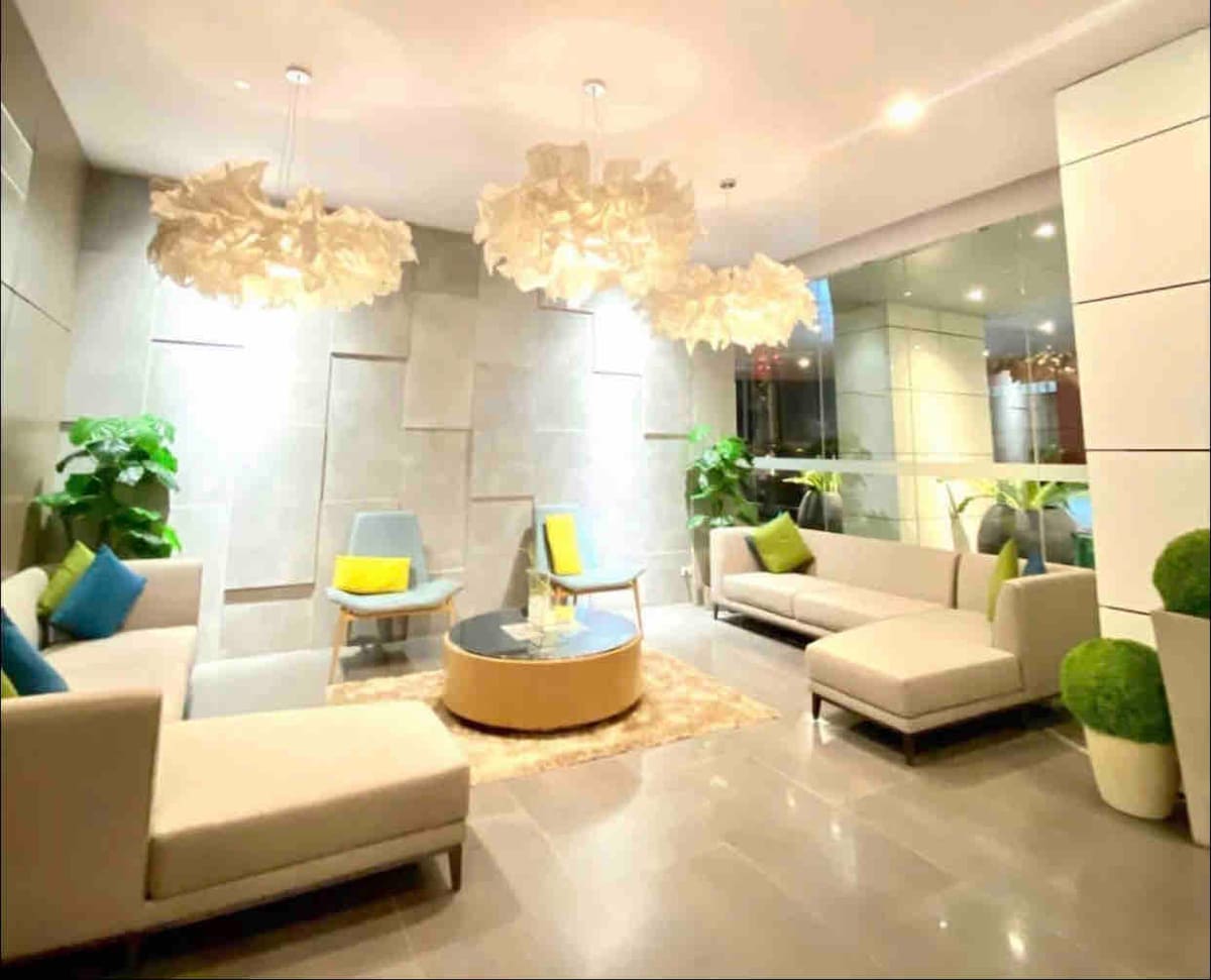 A modern lounge area is featured with soft seating arrangements and decorative plants. Two uniquely shaped light fixtures hang from the ceiling, providing soft illumination. A round coffee table sits at the center, accompanied by a large area rug that adds warmth to the space.