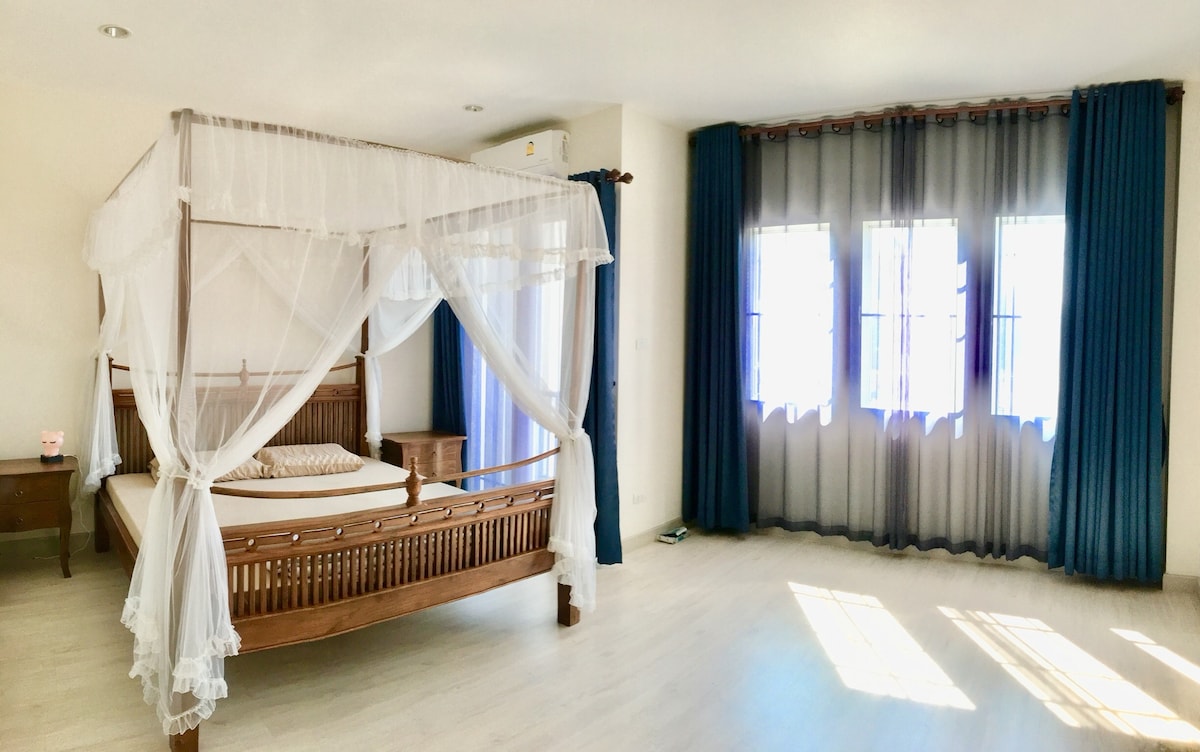 A spacious bedroom features a wooden four-poster bed with a delicate canopy. Natural light filters through large windows adorned with blue and sheer drapes, illuminating the light-colored flooring. A bedside table is positioned next to the bed, enhancing the room's functionality.
