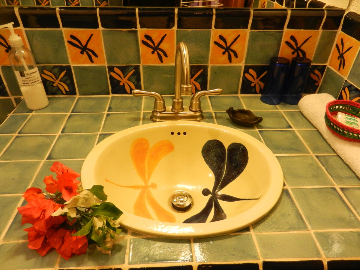 A brightly colored bathroom sink features intricate dragonfly designs in orange and black on a cream surface. The tiled backsplash showcases vibrant geometric patterns, while decorative towels, a soap dispenser, and flowers nearby add a touch of charm.