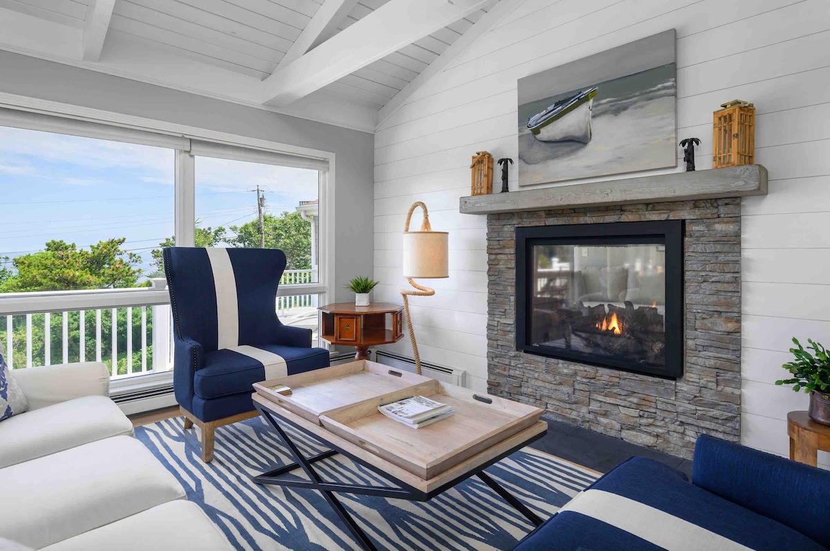 A living room features comfortable seating with a blue and white color scheme, complemented by a stone fireplace. A wooden coffee table sits centrally on a striped area rug. Large windows frame views of the outdoors, allowing natural light to illuminate the space.