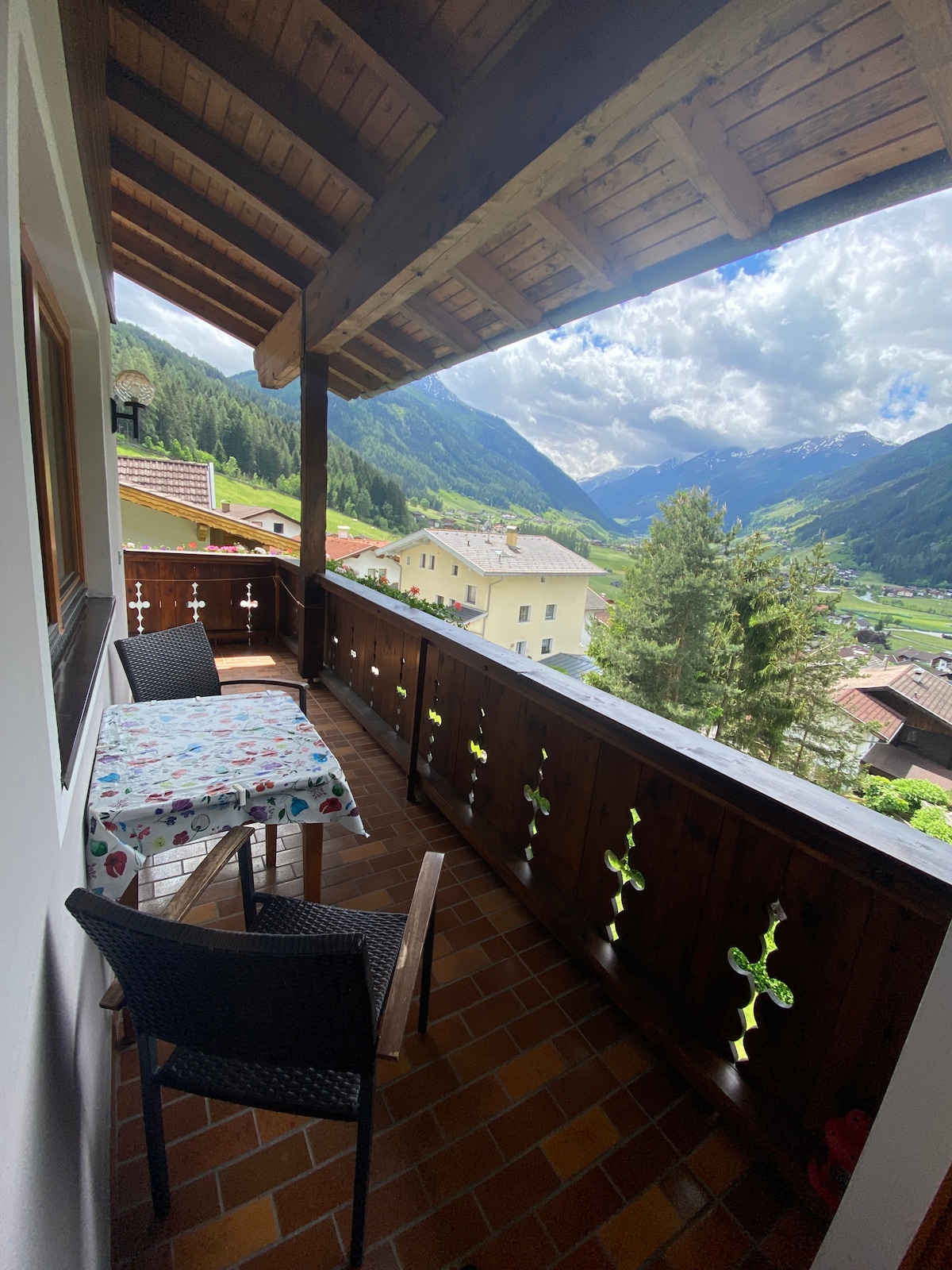 A balcony features wooden railings with decorative cutouts, offering scenic mountain views. A small table is set with a floral tablecloth, accompanied by two chairs. The surrounding landscape includes lush greenery and distant mountains under a partly cloudy sky.