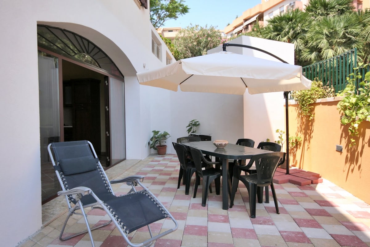 A spacious outdoor area is presented with a circular table and six black chairs, shaded by a large umbrella. A reclining lounge chair is positioned nearby, and potted plants add greenery to the space, enhancing the welcoming ambiance of the patio.