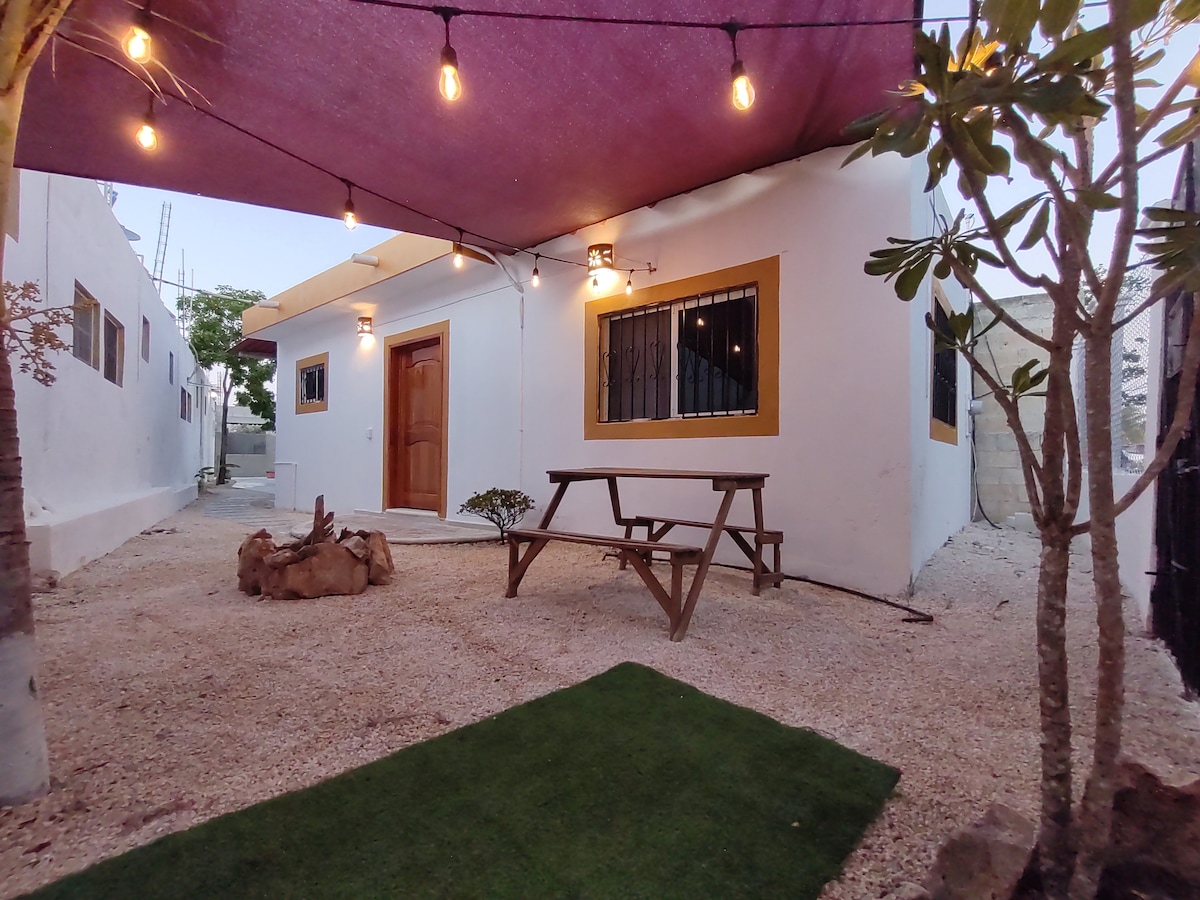 A charming patio area is showcased, featuring a rustic picnic table surrounded by gravel and greenery. Soft outdoor lighting hangs above, enhancing the evening ambiance. A wooden door leads into the house, with a small tree and plants adding natural elements to the space.