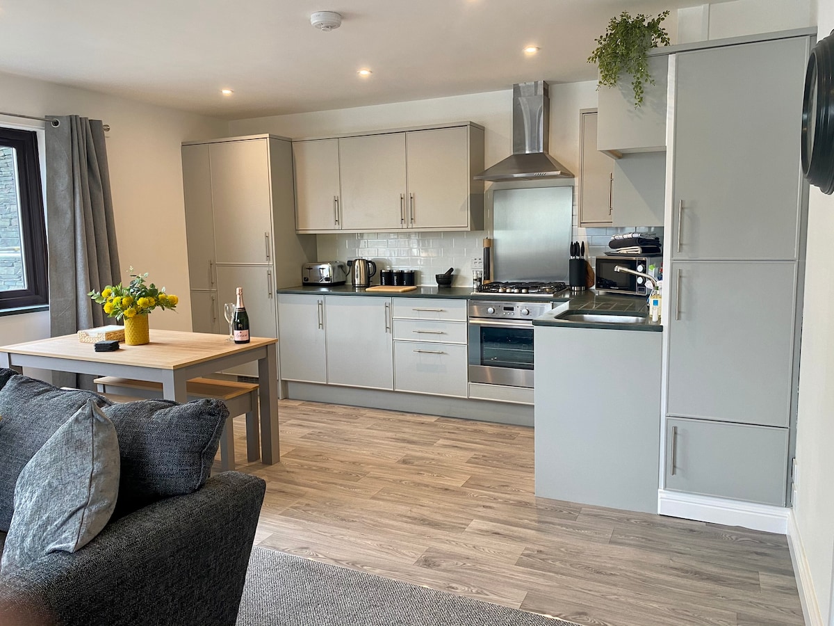 The kitchen and living area feature modern cabinetry and appliances. A dining table with seating for four is positioned near the compact kitchen. Two gray sofas offer comfortable seating, and a small bouquet is centered on the table, creating a welcoming atmosphere.