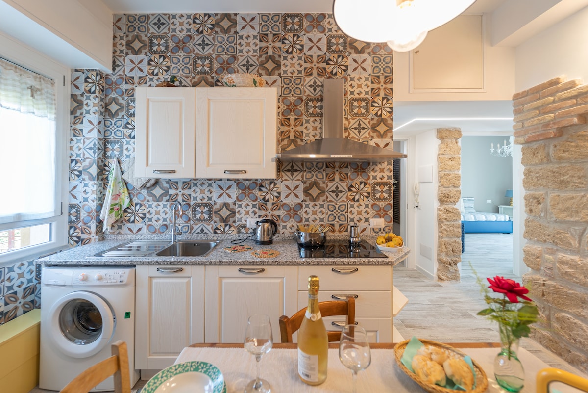 A well-equipped kitchen is featured, showcasing patterned tiles and a modern cooking area. Appliances include a washing machine and stove. A dining table is set for four, adorned with a flower centerpiece. Natural light filters through the window, illuminating the inviting space.