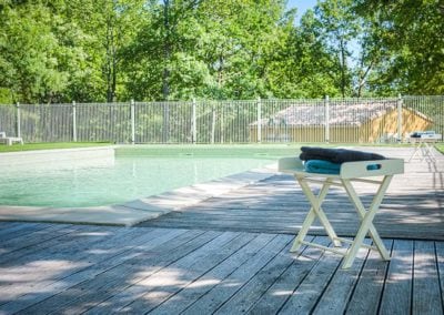 A swimming pool is surrounded by lush greenery, with a wooden deck leading to the water's edge. A folding tray table holds neatly stacked towels, positioned for convenience. A secure fence encircles the area, ensuring privacy and safety.