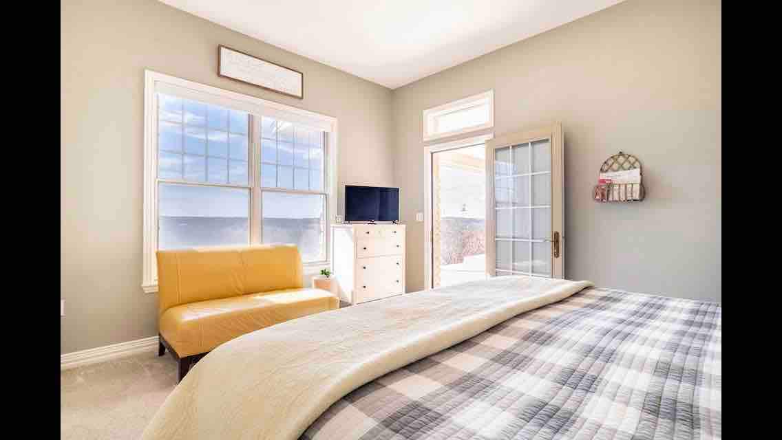A light-filled king suite features large windows with serene views and a comfortable seating area. A cozy bed is covered with a plaid blanket, while a small television and dresser are visible in the room. Access to a private patio is available through glass doors.