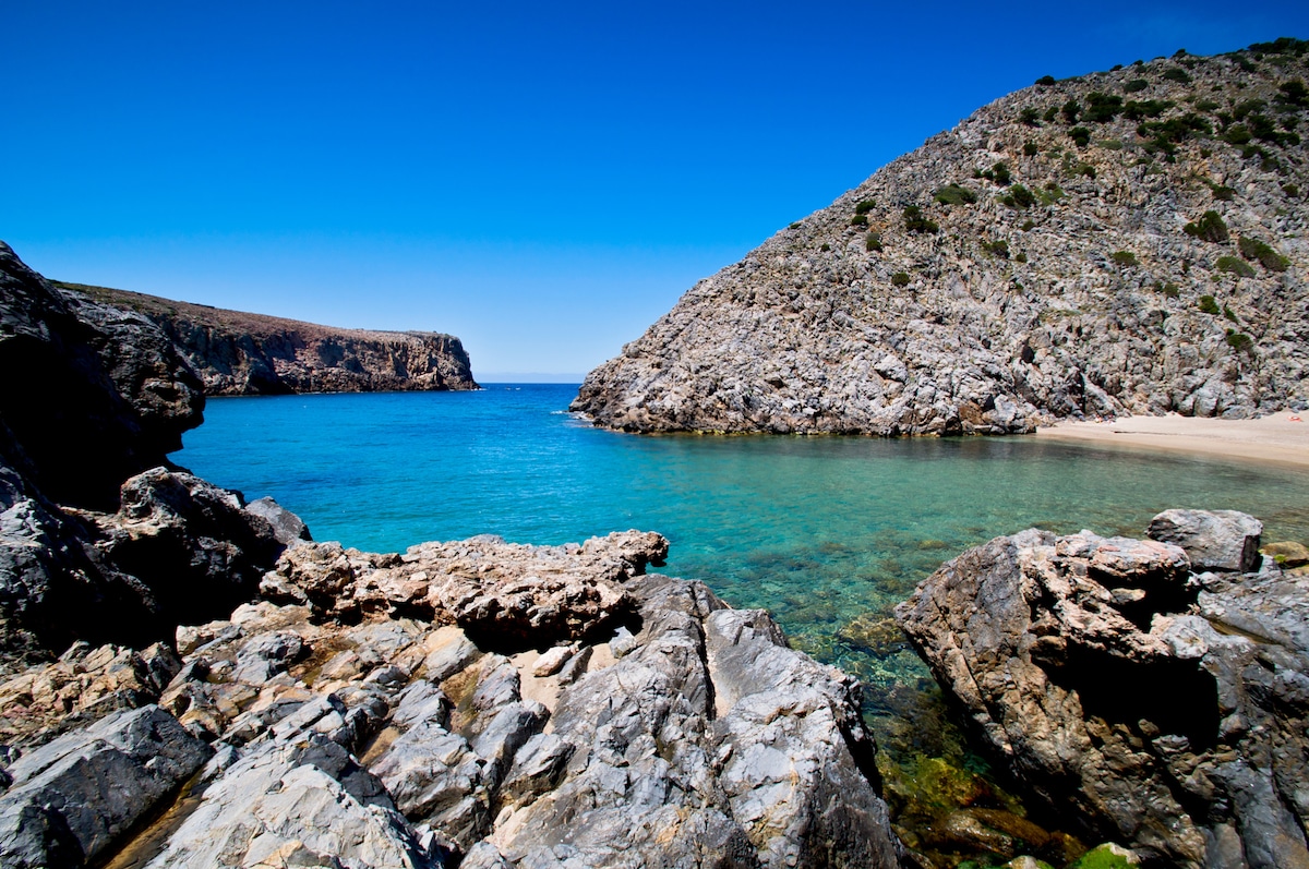 A serene coastal view features calm turquoise waters gently lapping against rocky shores. The landscape is framed by rugged cliffs, creating a secluded inlet. The clear sky above is bright, enhancing the tranquil atmosphere of this natural setting.