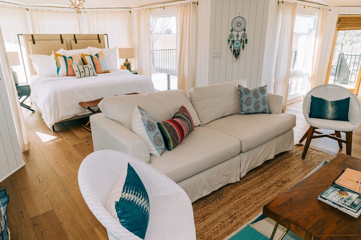 A cozy living area features a light-colored sectional sofa adorned with colorful pillows. A wooden coffee table sits on a natural fiber rug, while soft curtains frame large windows that provide ample natural light. The spacious bed with decorative pillows is visible in the background.