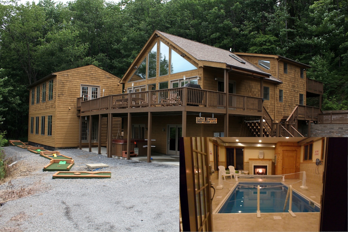 The exterior of a spacious wooden house features large windows and multiple entrances. An expansive deck surrounds the building, with outdoor mini golf areas visible. Inset is a heated indoor pool area, complete with a fireplace, contributing to a relaxing atmosphere.