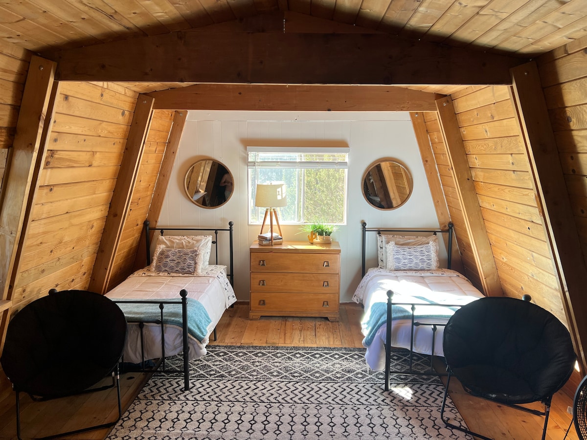 An A-frame loft space features two individual beds, each dressed in light bedding. A wooden dresser sits between the beds, accompanied by a lamp and a small plant. Two circular mirrors are mounted on the slanted walls, enhancing the cabin's wooden aesthetic.