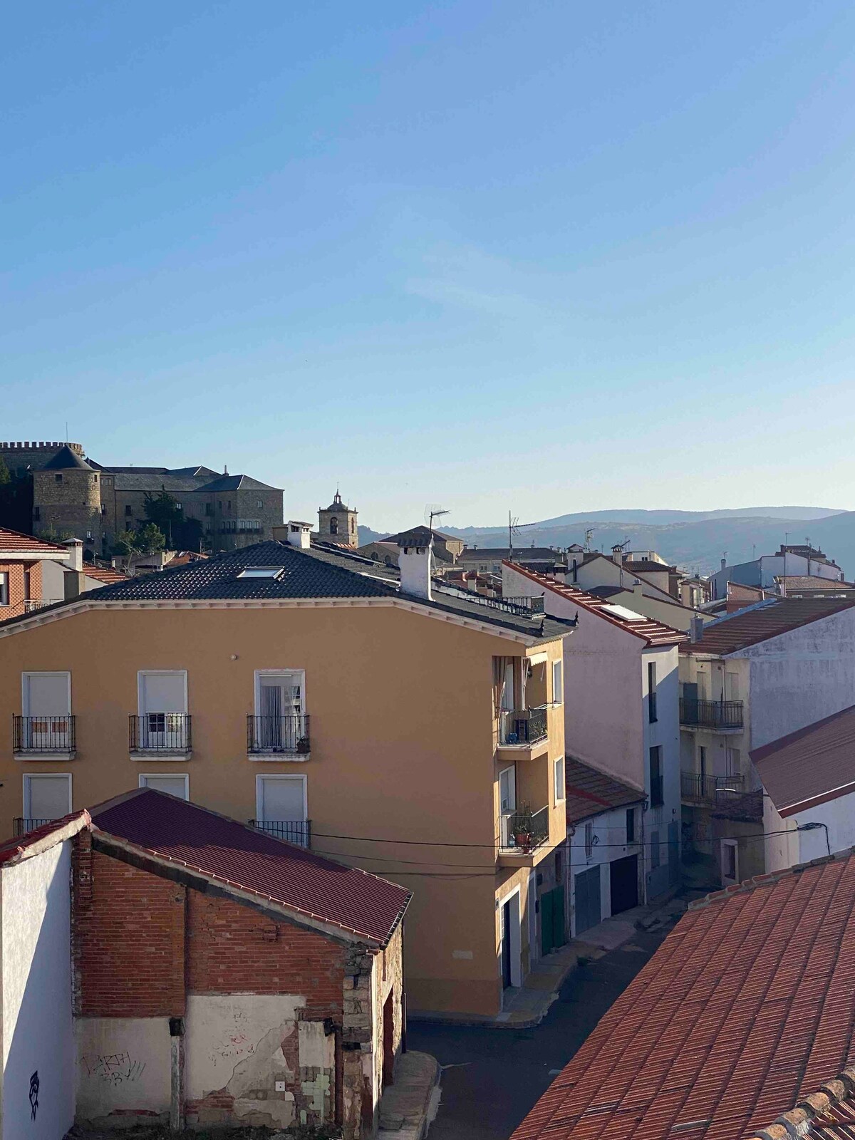 A panoramic view is captured from the apartment window, displaying charming houses with various colored facades and tiled roofs. In the background, the silhouette of the Castillo de Magalia can be seen against a clear blue sky, enhancing the picturesque landscape of the town.