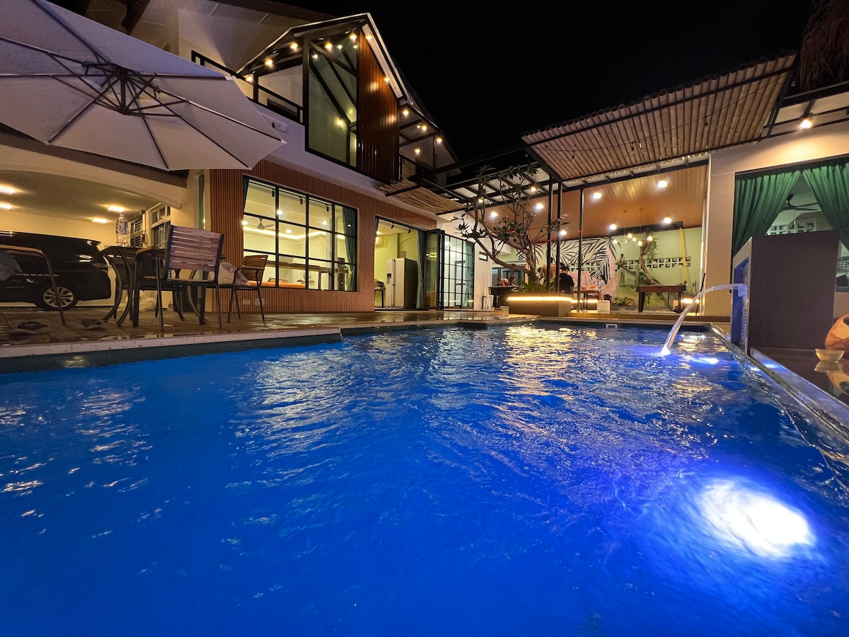 The illuminated pool reflects vibrant blue tones under the evening sky. Surrounding features include a modern architecture with large glass windows, outdoor seating arrangements, and a softly lit patio area, creating a serene ambiance for relaxation.