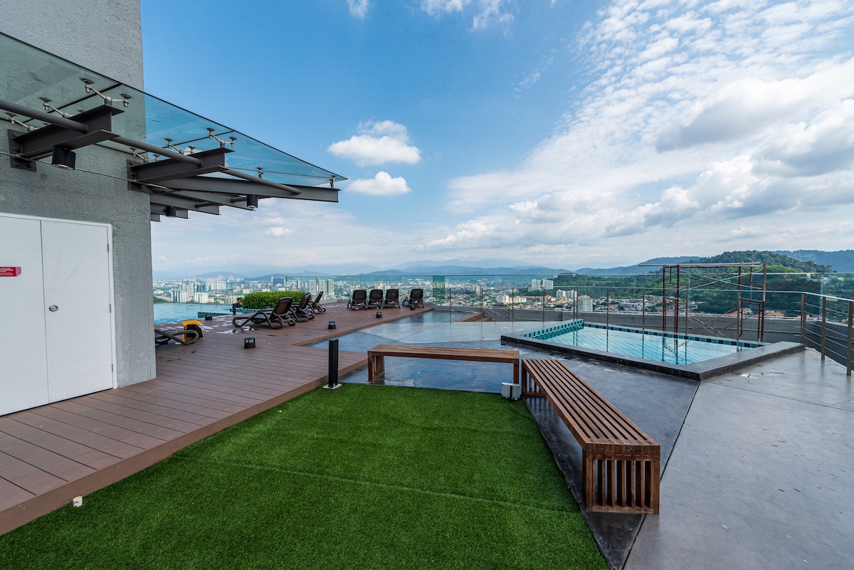 An outdoor rooftop area presents a poolside view overlooking the cityscape. A small pool reflects the sky, while loungers are positioned for sunbathing. A grassy section adds greenery, and a wooden bench offers a place to relax, with glass railings ensuring safety and an unobstructed view.