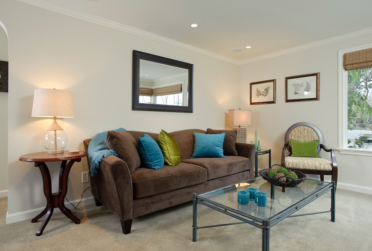 A cozy living area features a brown couch adorned with green and blue cushions. A glass coffee table sits in the center, surrounded by tasteful decor. Soft lighting from a nearby lamp creates a warm ambiance, while artwork is displayed on the walls.