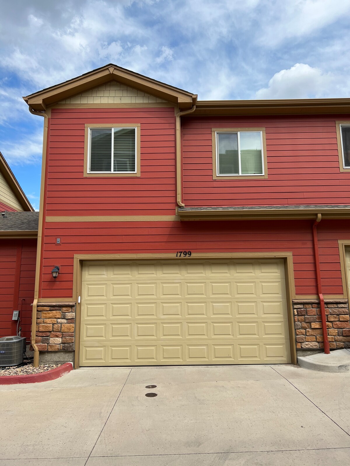 A two-story townhome is shown, featuring a red exterior with contrasting beige accents. A stone detail is visible at the base, and a garage door is situated on the lower level. Windows are present on both levels, allowing natural light to enter.