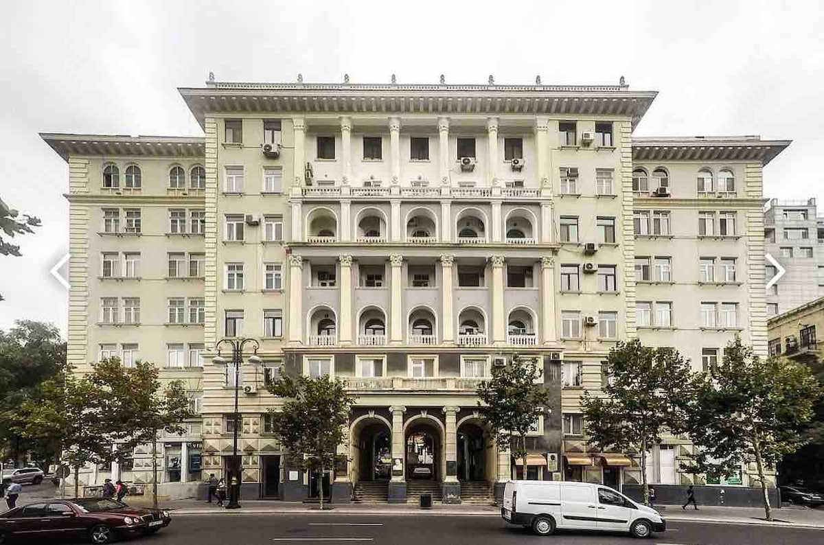 A historic building with an elegant facade showcases multiple balconies and large arched windows. The structure is framed by trees and street-level activity, highlighting its vibrant urban setting. Air conditioning units are visible, indicating a comfortable indoor environment.
