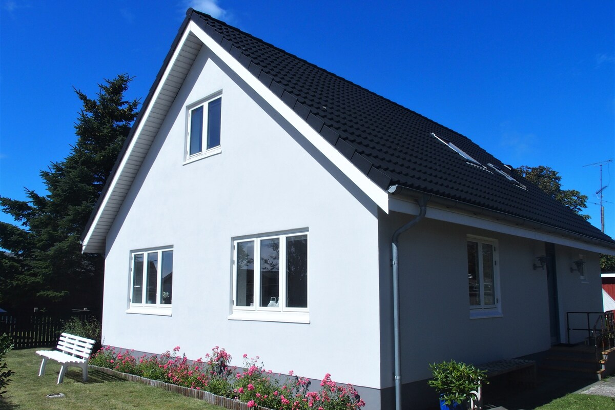 A white, two-story house is depicted with a sloped black roof and multiple windows reflecting daylight. Surrounding the house, a well-maintained garden showcases blooming flowers and a seating area, contributing to a peaceful outdoor setting.