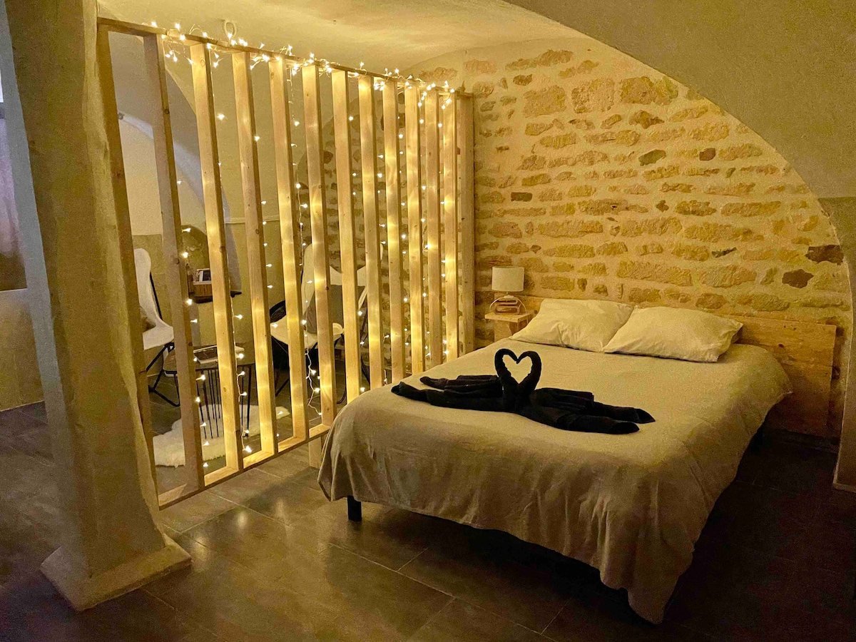 A spacious bed is positioned against a textured stone wall, with soft bedding and decorative towels arranged neatly. A wooden partition adorned with warm string lights creates a cozy separation from the adjacent area, which includes a small workspace and a chair.