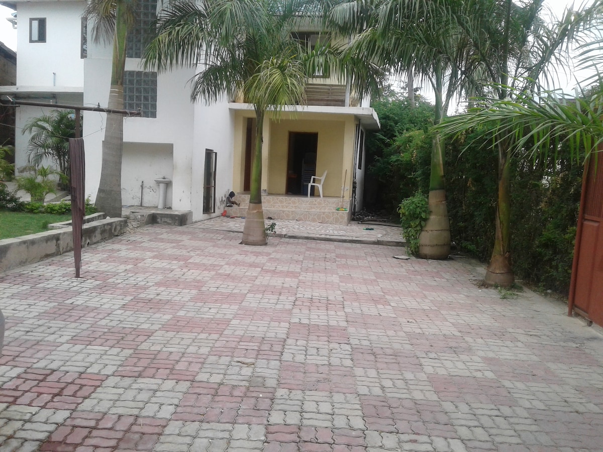 A spacious outdoor area is surrounded by tall palm trees, featuring a patterned stone floor that leads to the main entrance. The entrance is framed by a porch area, with a chair visible just inside. The site offers ample room for movement and relaxation.