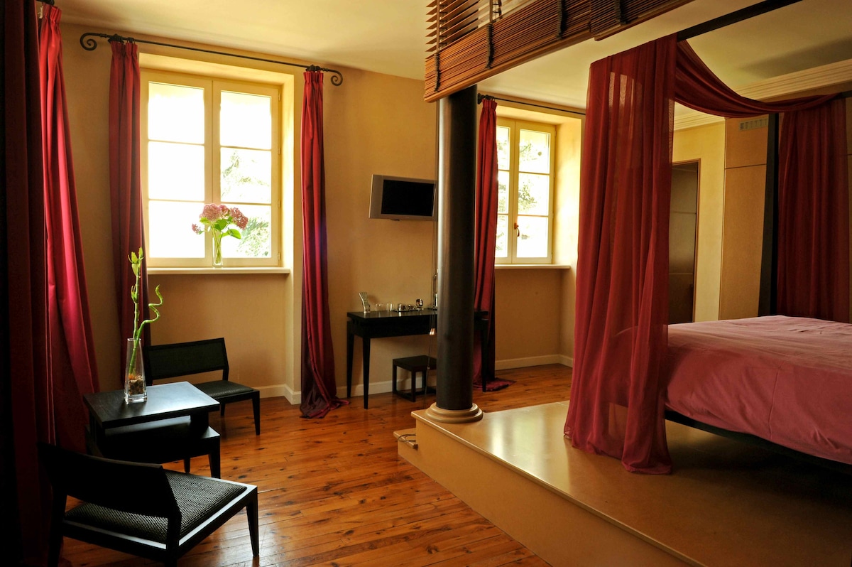 The room is designed with soft colors and warm wood flooring. A spacious bed is framed by sheer, flowing drapes. Two seating areas feature simple chairs with a small table beside a decorative vase. Windows allow natural light to fill the space.