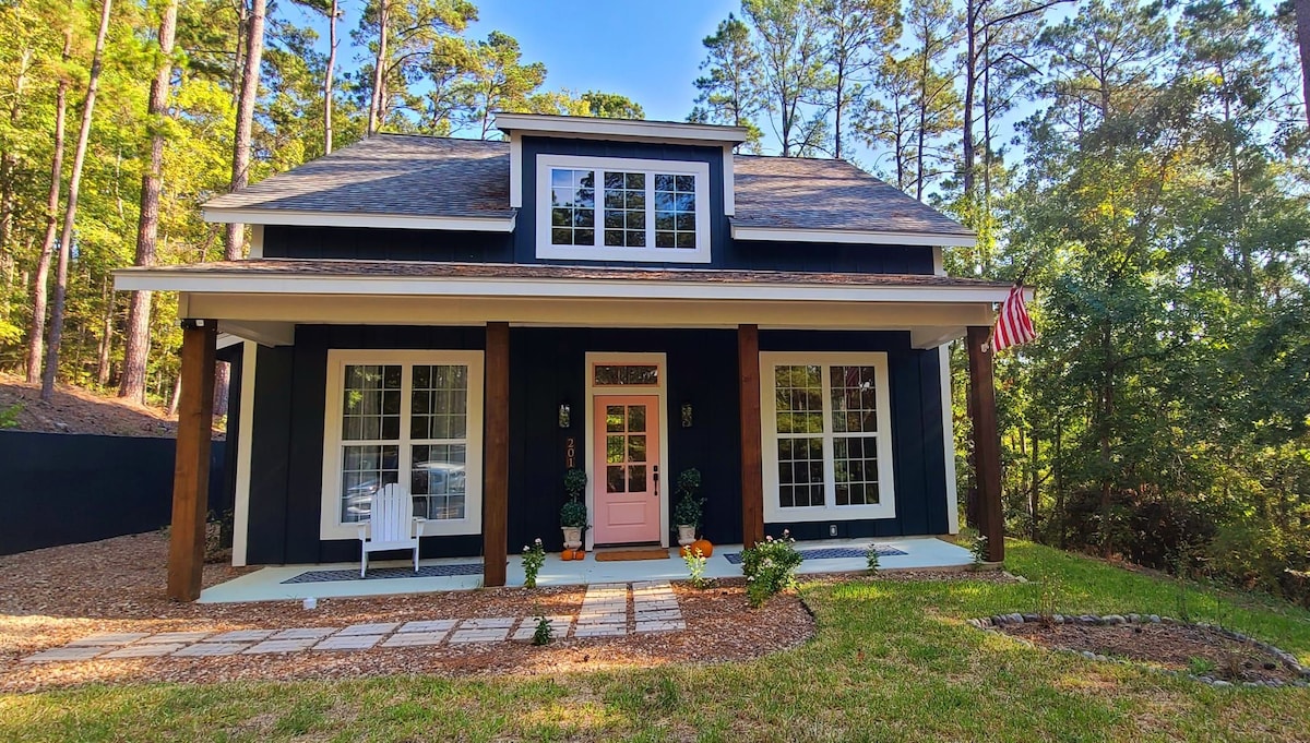 The exterior view of a two-story cottage is presented, showcasing a dark blue facade accented by white trim. A welcoming pink front door is flanked by windows and plants. Stone pathways lead to the entrance, and tall pine trees provide a natural backdrop.
