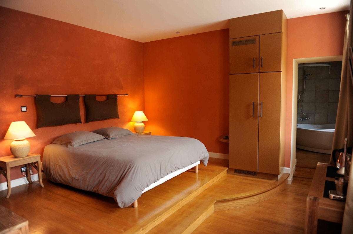 A cozy bedroom features a spacious bed with neutral bedding, positioned on a raised platform. Soft lighting from two bedside lamps creates a welcoming ambience. A built-in wardrobe is visible, and a hint of an elegant bathtub can be seen through an open doorway.