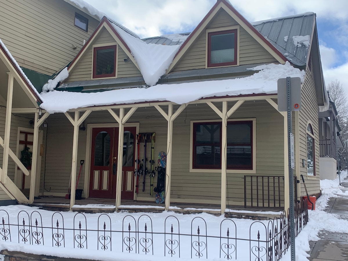 A charming two-story cottage is shown, featuring a sloped roof and a welcoming porch supported by decorative columns. Equipment for skiing is visible against the wall, and a well-kept pathway leads to the entrance, surrounded by fresh snow.