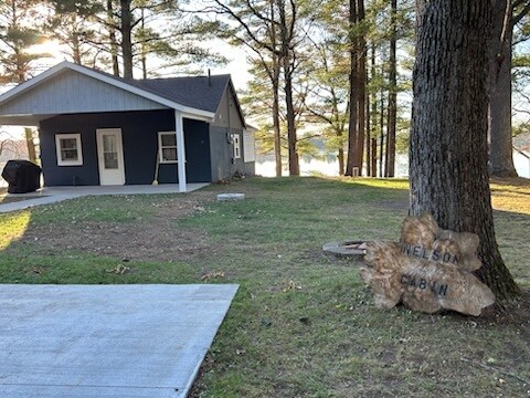 The cabin exterior is set amidst tall trees, with a clear view of the lake. A natural stone marker displays 'Nelson Cabin' prominently. The patio area in front offers ample space, complemented by a grill placed nearby.