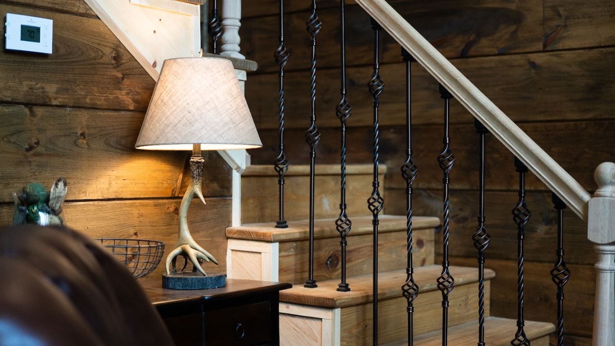 The image shows a staircase crafted from light-colored wood, with wrought iron balusters featuring intricate detailing. A warm lamp with a textured shade illuminates the surrounding area, complementing the natural wood paneling that provides a cozy atmosphere.