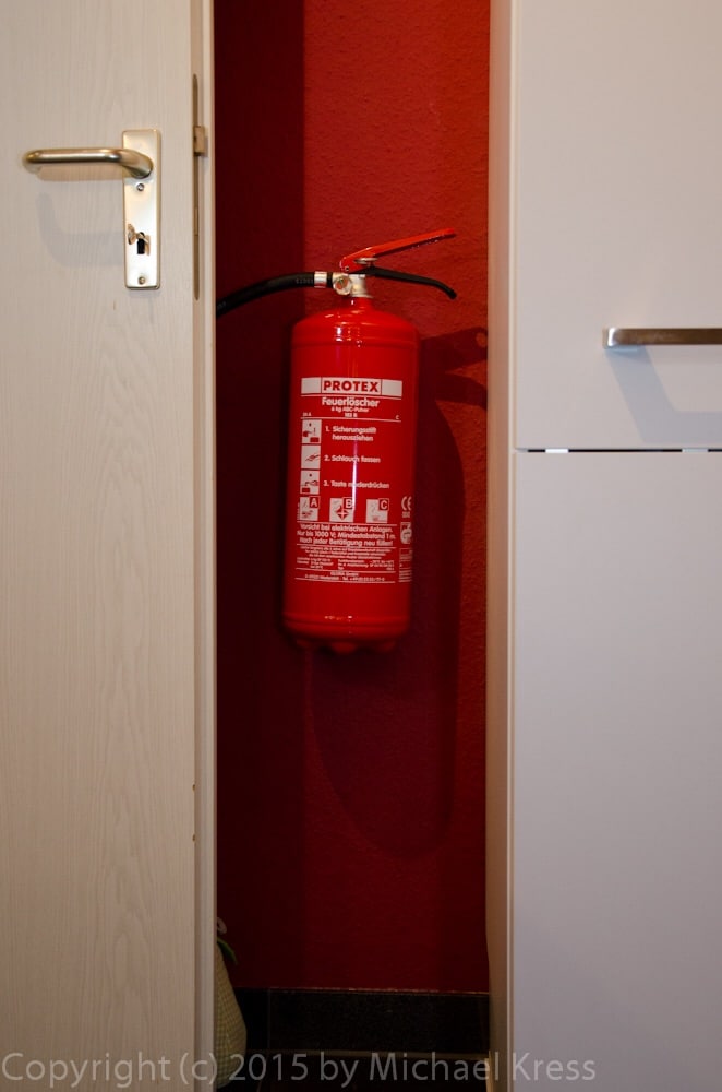 A fire extinguisher is mounted on the wall, easily accessible in case of an emergency. It is housed in a narrow space between two cabinets, with a contrasting red color against the backdrop of a warm red wall.