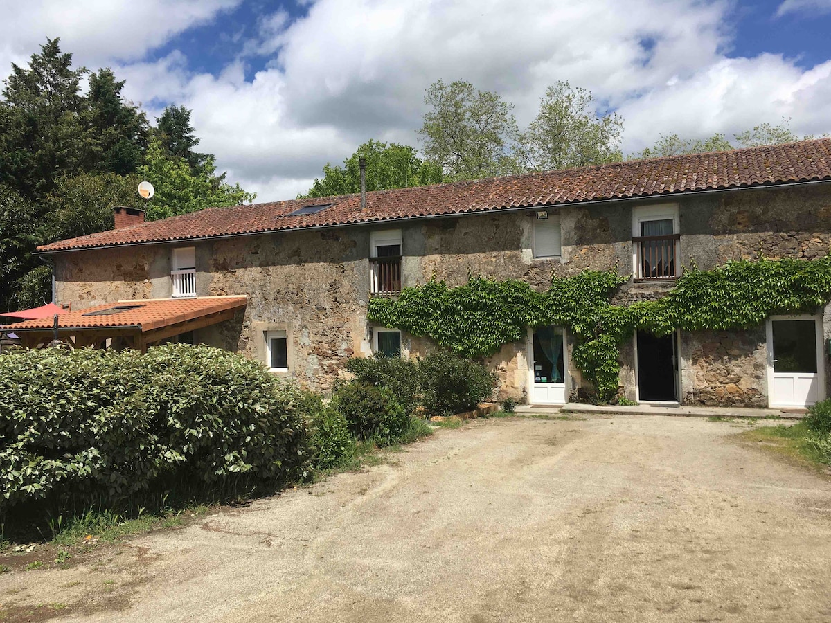An old stone building is framed by greenery, showcasing a rustic façade with climbing plants. The structure has several windows and a covered entrance that leads to a gravel path. The surrounding landscape includes trees, enhancing the tranquil rural setting.