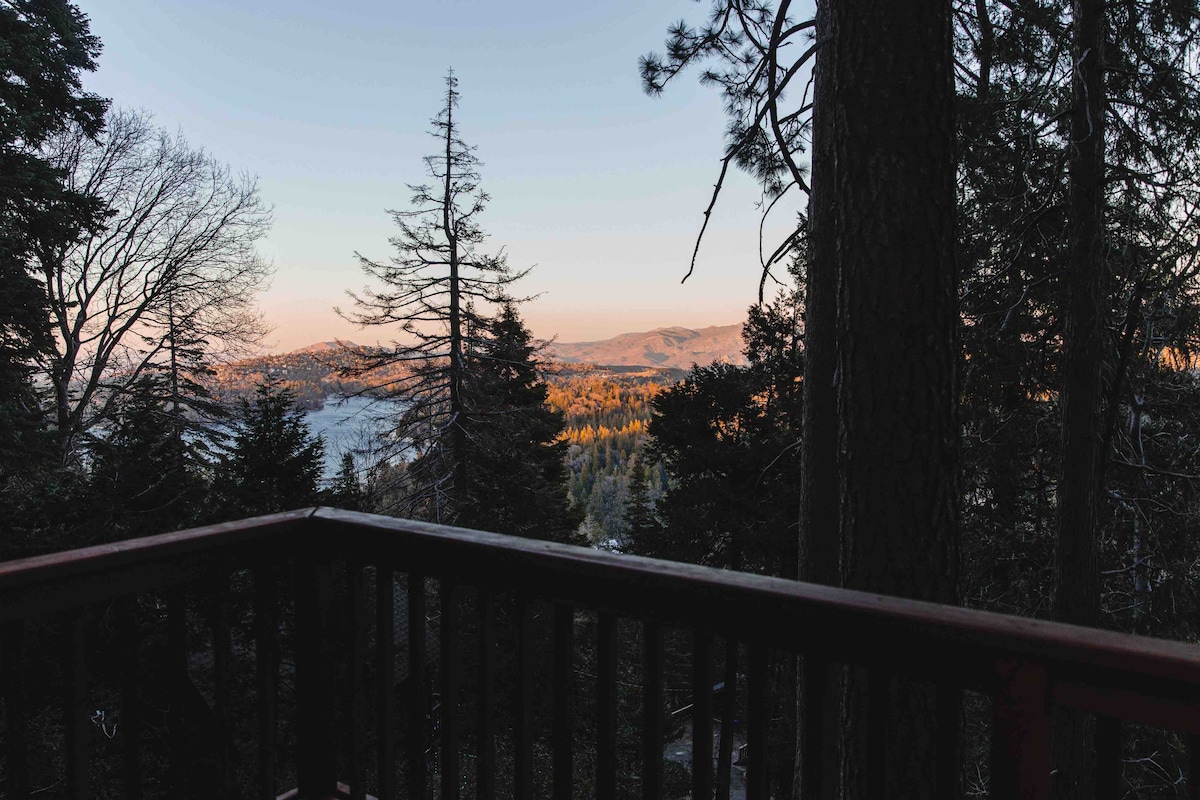 A wooden railing encloses a balcony, offering a clear view of the surrounding landscape. The distant mountains are bathed in soft morning light, while tall trees frame the scene. The tranquility of nature is evident as the sun rises over a forested area.