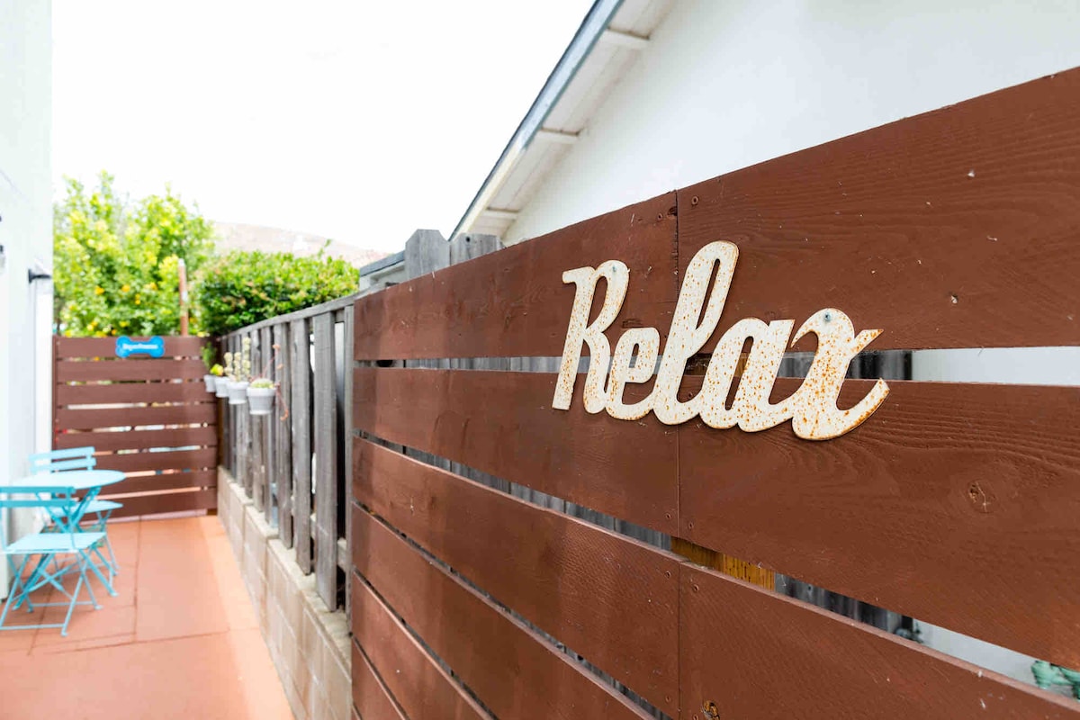 A wooden fence lined with planters creates a peaceful outdoor space. A 'Relax' sign adorns the fence, complementing the bright blue chairs and table positioned nearby. Lush greenery can be seen in the background, adding to the inviting ambiance.