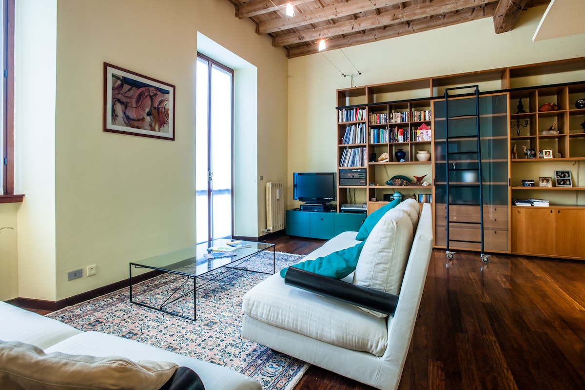 A spacious living room features large windows that allow natural light to fill the space. Stylish, minimalist furniture includes a comfortable sofa, a glass coffee table, and a sleek television unit. A wooden shelving unit displays books and decorative items, enhancing the modern aesthetic.