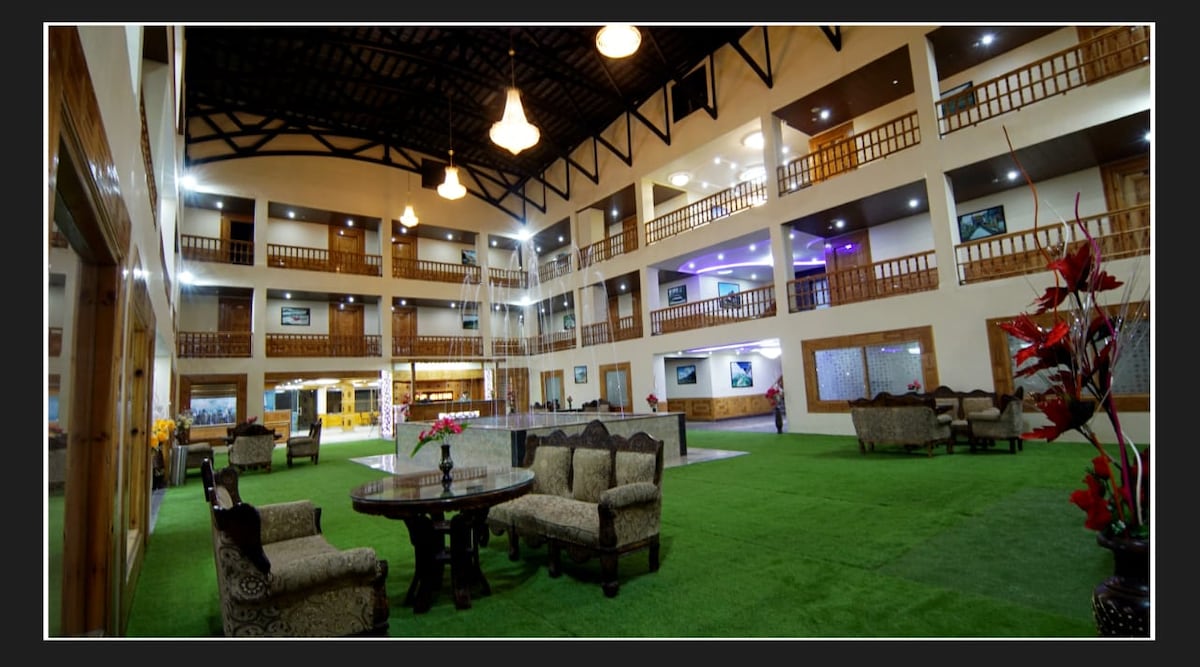 The spacious lobby features a high ceiling with wooden beams, creating an open atmosphere. Comfortable seating arrangements include multiple chairs and round tables, surrounded by lush green carpeting. A central musical fountain adds a focal point, with soft lighting enhancing the overall ambiance.
