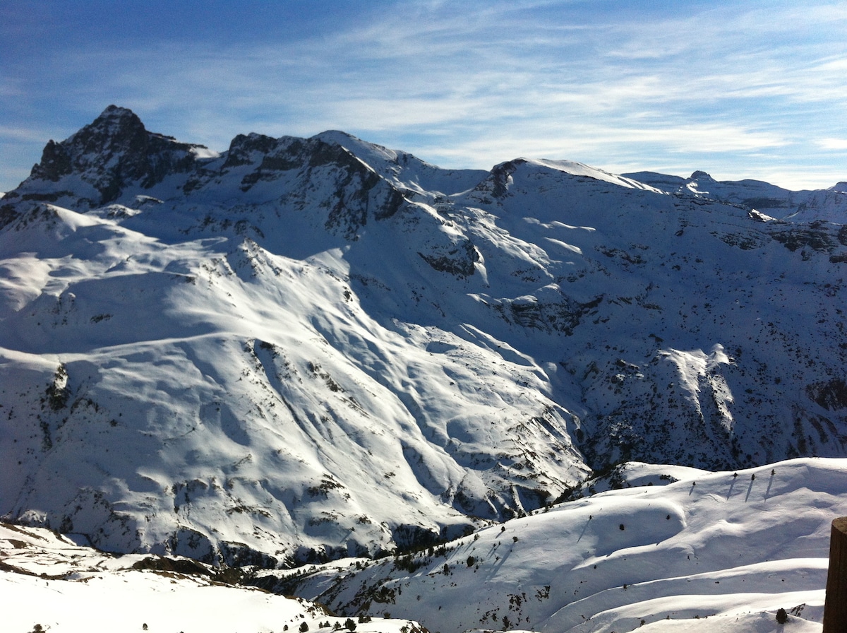 Snow-covered mountains rise majestically against a clear blue sky. The vast landscape is marked by rolling hills and peaks, creating a serene winter tableau. The light reflects off the snow, adding a pristine quality to the scene.