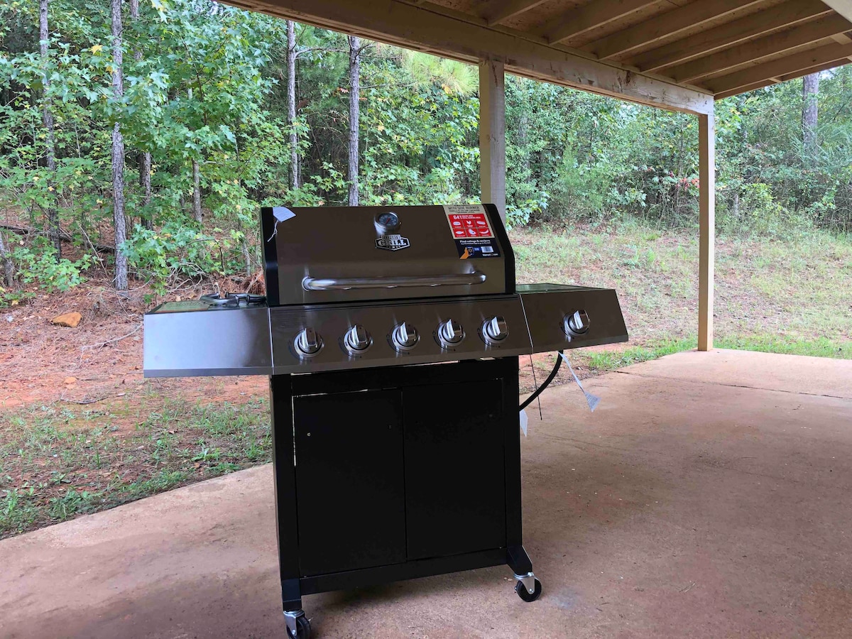 A stainless steel gas grill is positioned under a covered patio, featuring multiple burners and a sleek black cabinet below. The surrounding area includes dense trees and natural greenery, offering a serene outdoor cooking space.