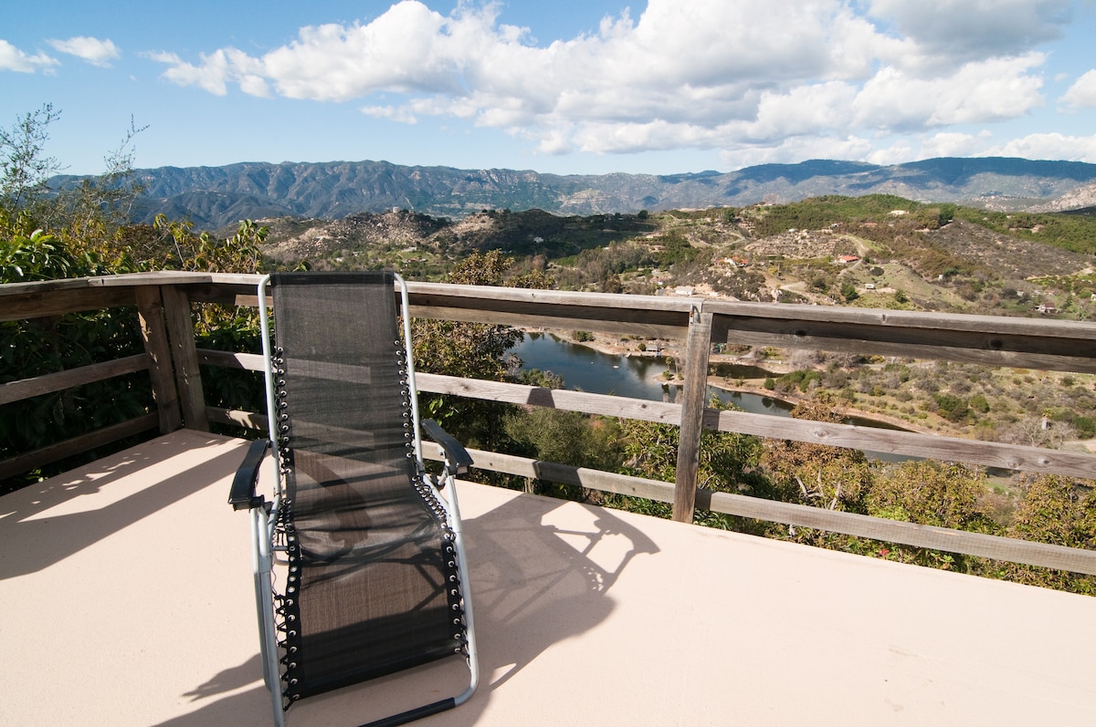 An outdoor deck is presented with a single black reclining lounge chair positioned against a wooden railing. In the background, rolling hills provide a panoramic view of the landscape, including a glimpse of a tranquil lake under a partly cloudy sky.