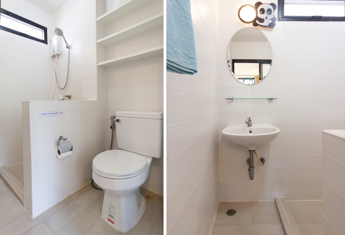 A clean and minimalist bathroom is shown, featuring a modern shower with an adjustable head positioned above the sleek tiled space. A compact toilet is situated beside a small sink with a circular mirror above it. Shelving is offered for storage, contributing to the functional design.
