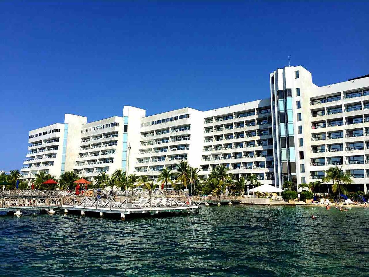 The beachfront building is seen from the water, showcasing a modern architectural design with multiple balconies. Lush palm trees surround the area, and a dock extends into the clear blue water, providing easy access to the shoreline and amenities.