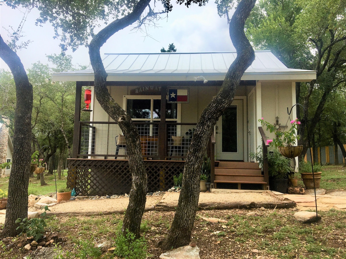 A charming cottage is framed by two trees in the foreground. The front porch, equipped with a railing, showcases a welcoming entryway adorned with a Texas flag and lanterns. Lush greenery surrounds the property, enhancing the rural ambiance.