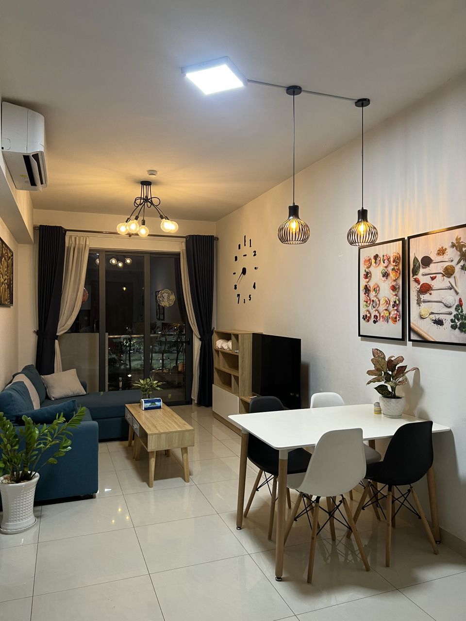 A modern living space features a blue sofa, a light wood coffee table, and a dining area with white chairs around a rectangular table. Decorative artwork adorns the walls, while a balcony can be glimpsed through large glass doors, providing natural light to the room.