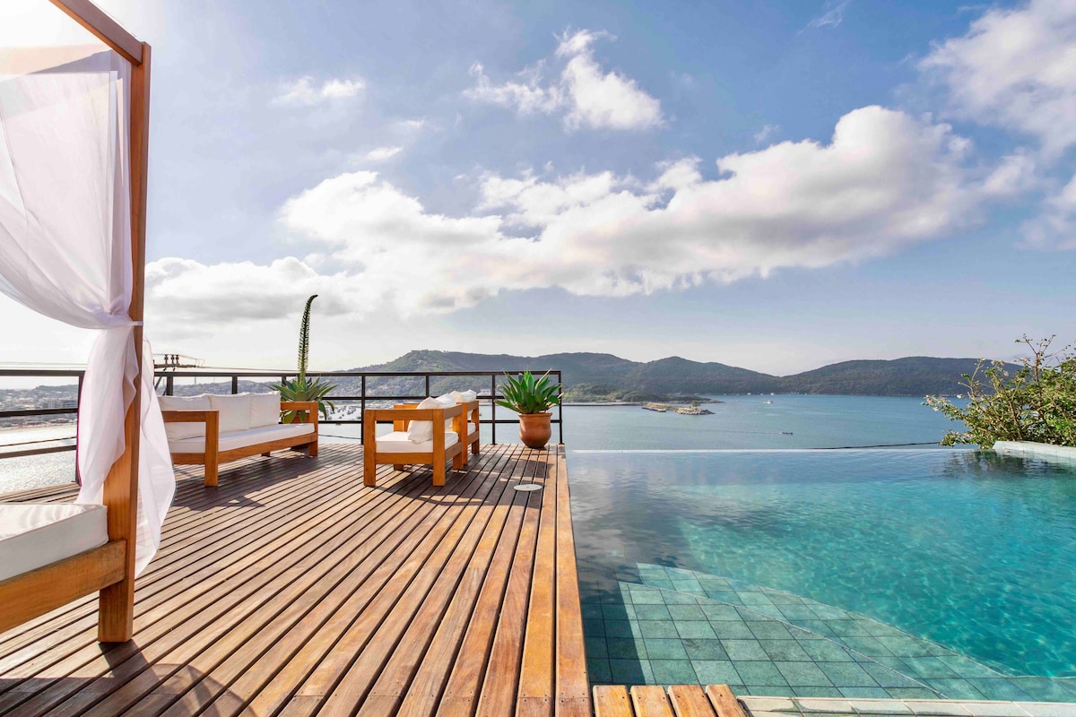 An infinity pool extends towards the horizon, framed by wooden decking and lounge chairs adorned with soft cushions. Lush green plants accentuate the serene atmosphere, while panoramic views of mountains and sea are visible under a blue sky with soft clouds.