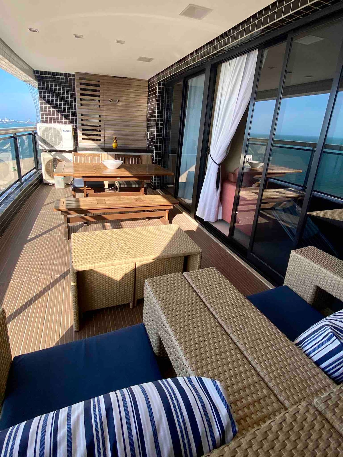 An outdoor balcony is presented, featuring seating arrangements with woven furniture and a wooden dining table. Large glass doors enhance the view of the sea, while a breezy atmosphere is suggested by the partially pulled sheer curtains. Decorative details include striped cushions on the seating.